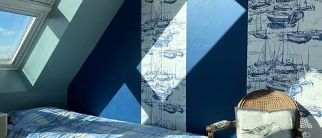A bedroom with a skylight, a bed with a blue checkered bedspread, a chair with a cushion, and wallpaper with a nautical theme.