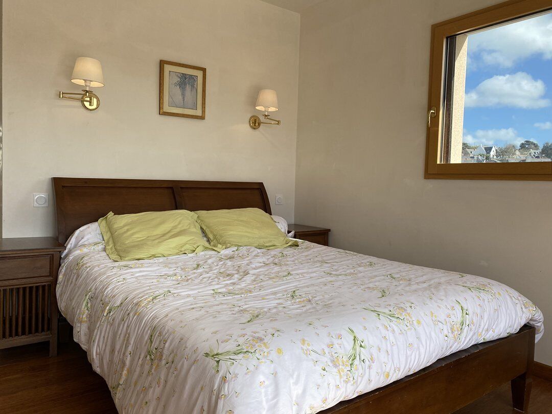 Un dormitorio con cama de madera, dos lámparas de pared, un cuadro enmarco y una ventana con vistas a edificios.