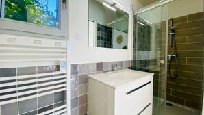 A modern bathroom with a glass shower enclosure, a white vanity with a mirror, and a view of greenery through a window. - Charming villa in a quiet location with courtyard (LE BOIS PLAGE EN RE)