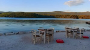 Um cenário à beira de um lago, com uma mesa e cadeiras em uma praia arenosa, com vista para um lago tranquilo e colinas arborizadas.