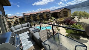 A balcony with outdoor furniture overlooking a swimming pool and a view of a hotel building with a pool area and beach chairs.