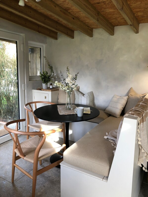 A cozy room with a wooden ceiling, a round table with chairs, a sofa, and a window with a view of greenery.