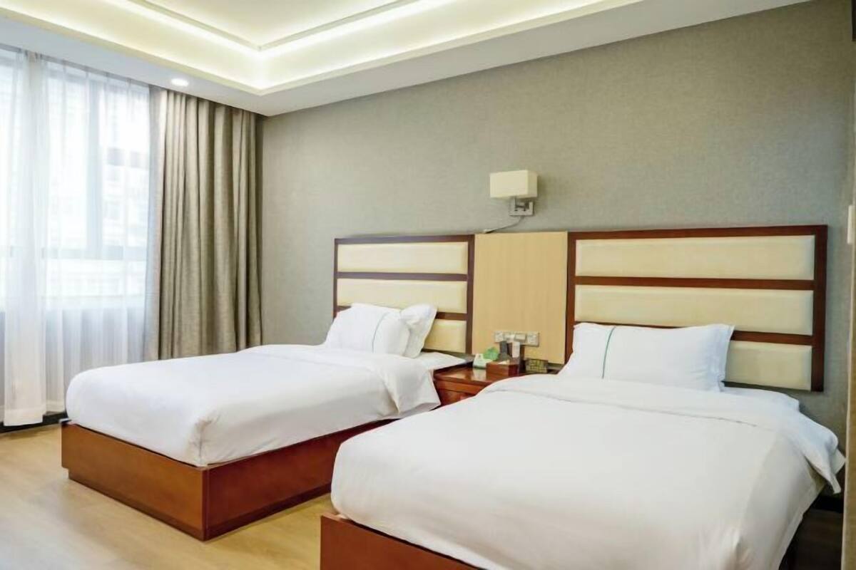 A hotel room with two beds, a nightstand, a lamp, and a view of the city through large windows.