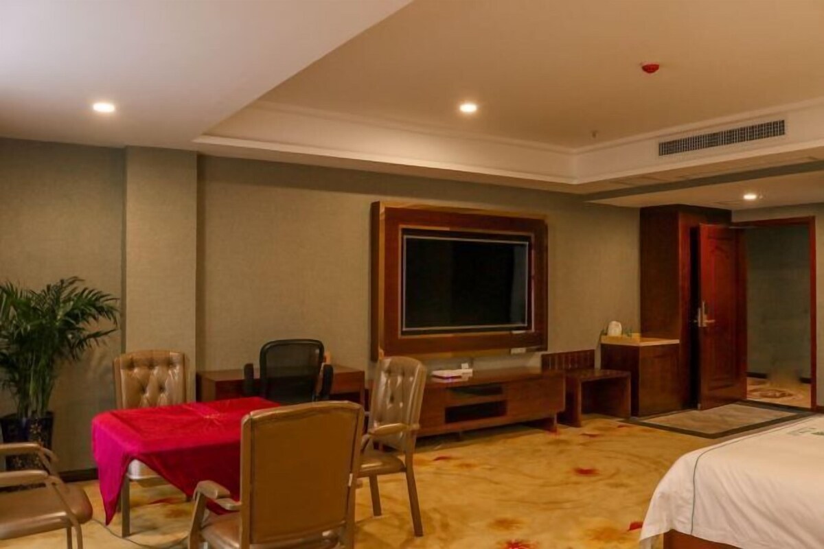 A hotel room with a bed, a desk with a computer, a TV, a plant, and a red tablecloth.