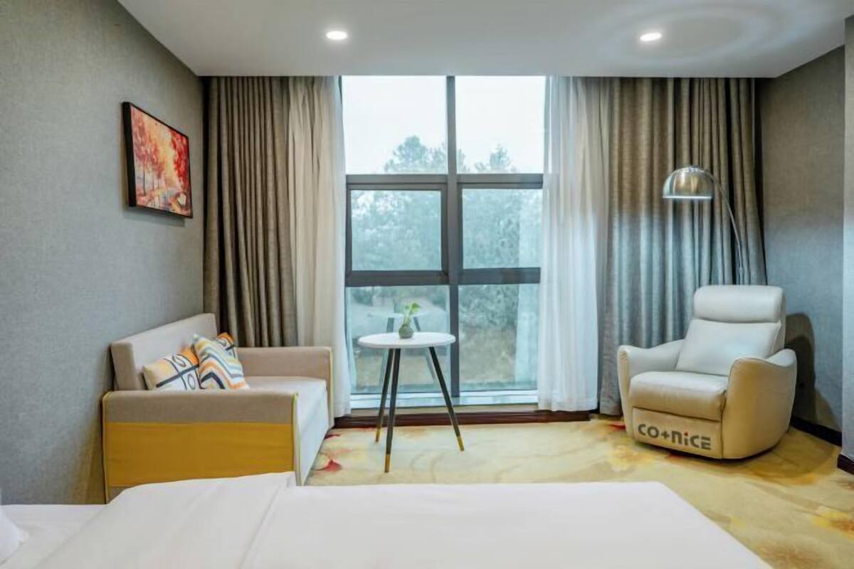 A hotel room with a bed, a sofa, a chair, a table, and a large window with curtains.