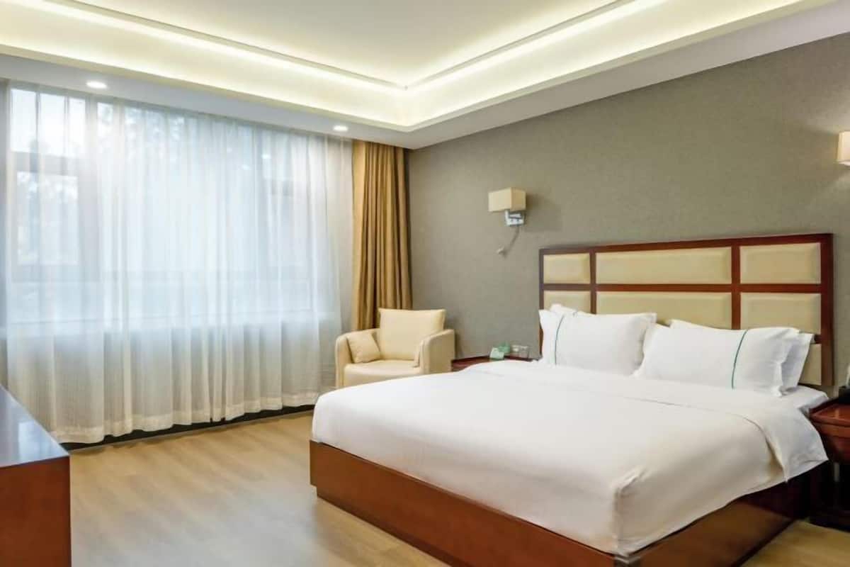 A hotel room with a large bed, a chair, a bedside table, and a window with sheer curtains.