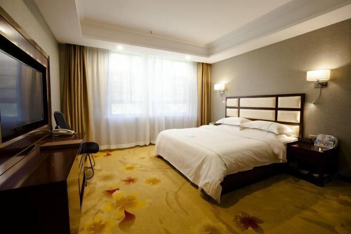 A hotel room with a bed, a desk with a television, a chair, and a nightstand with a lamp.