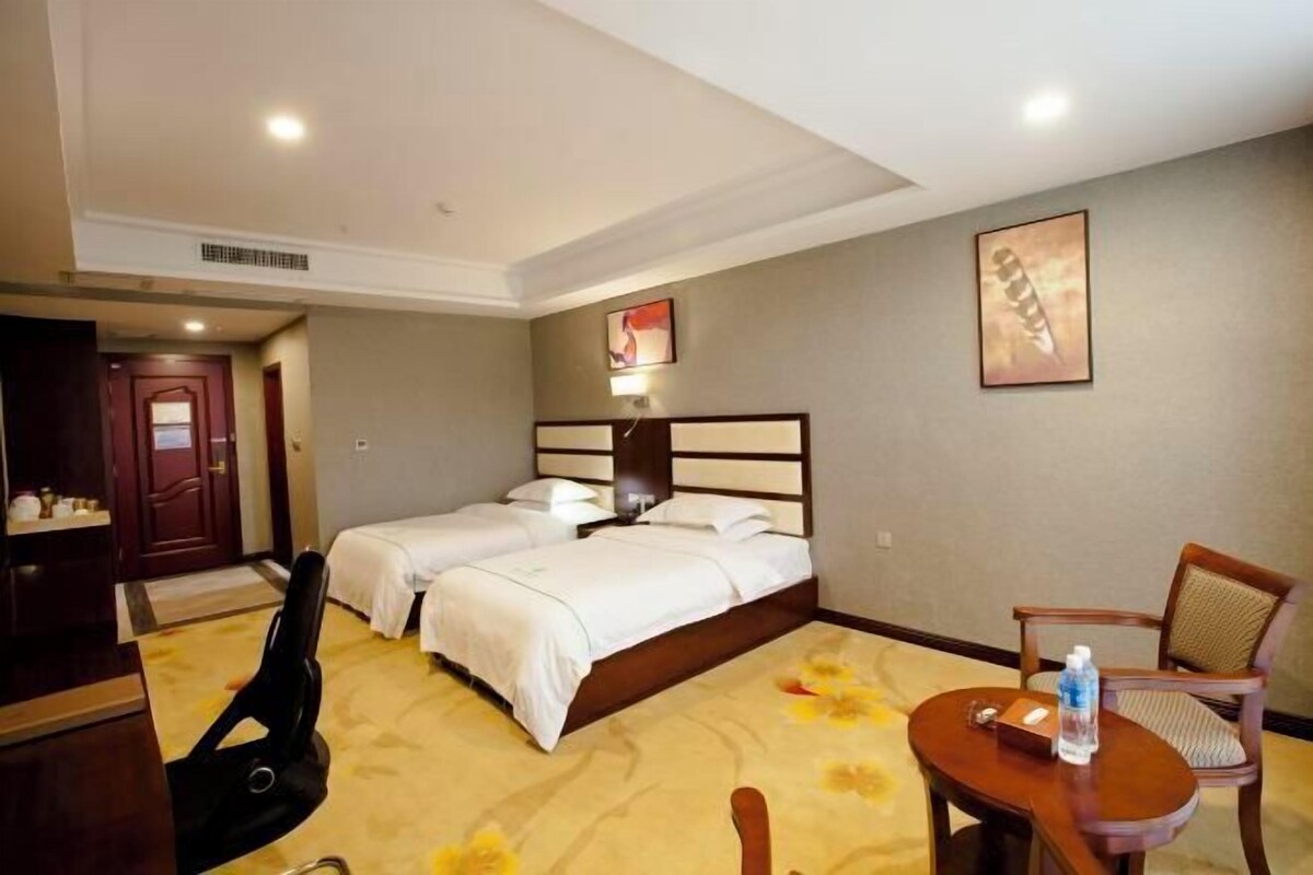 A hotel room with two beds, a desk, a chair, and a small table with a water bottle.