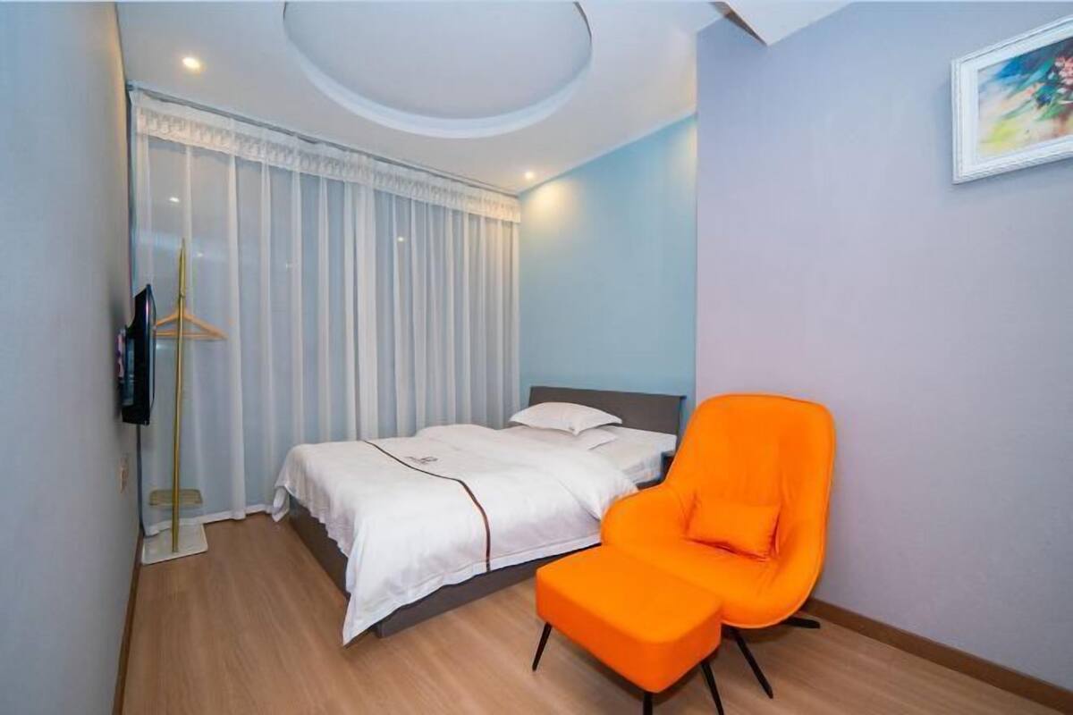 Habitació d'hotel amb un llit, una cadira taronja, una perxa i un quadre a la paret.