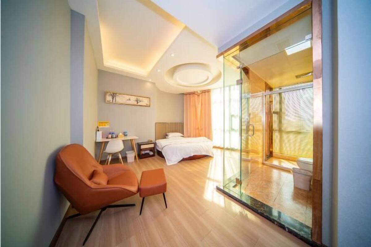 Habitació d'hotel amb llit, zona de seients amb una cadira i otomana, taula petita amb làmpada i gran porta de vidre que dona a un balcó.