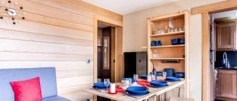 A compact living space with a wooden wall, a sofa with a red cushion, a dining table set with blue plates, and a kitchen area with wooden cabinets.