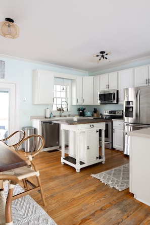 A modern kitchen with white cabinets, stainless steel appliances, and a wooden dining table. - Modern Boho style Folly beach house -3 blocks from the beach! (Folly Beach)