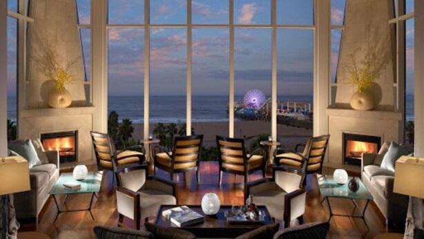 A spacious lounge with a large window overlooking a beach and a Ferris wheel, featuring comfortable seating and a fireplace. - La Plage Wellness (Santa Monica)