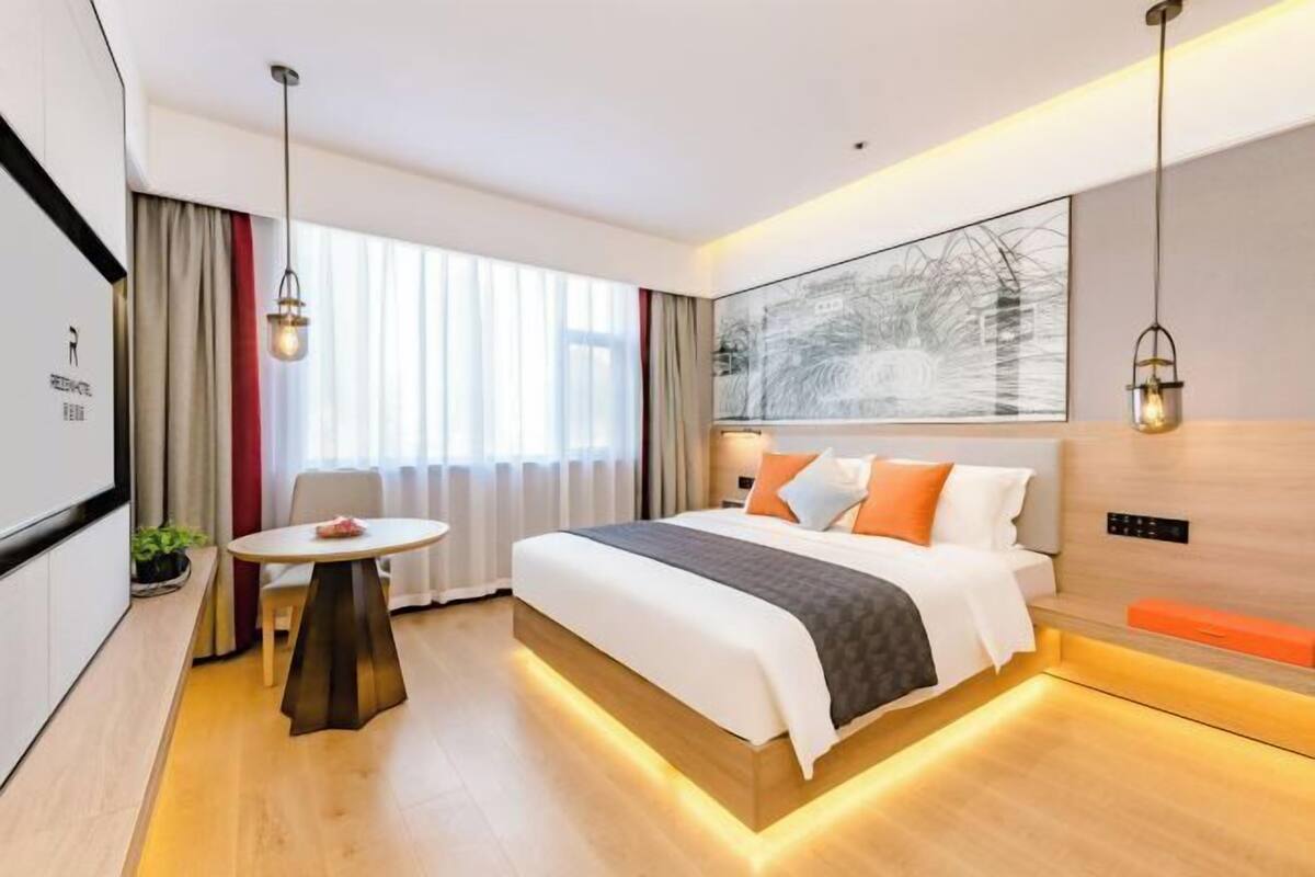 Ett modernt hotellrum med en säng, ett nattduksbord och ett stort fönster med gardiner.