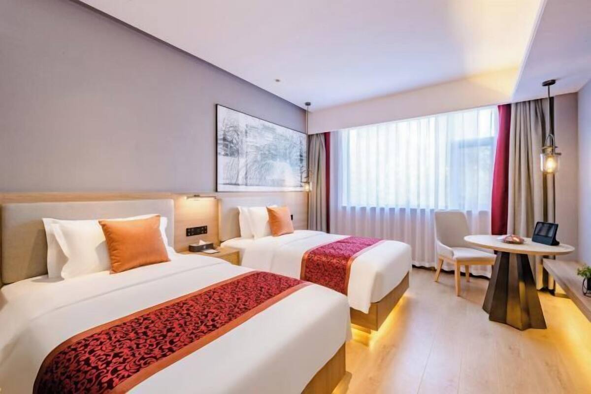 Ett hotellrum med två sängar, ett skrivbord, en stol, en lampa och ett fönster med gardiner.