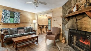A cozy living room with a wooden floor, a sofa with patterned cushions, a wooden coffee table, a patterned armchair, a fireplace with a stone surround, and a ceiling fan. -  Dog Friendly w/flat lot - Between Gatlinburg and Pigeon Forge! (Sevierville)