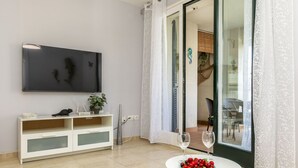 A modern living room with a flat-screen TV, a white console table with decorative items, a round table with a fruit bowl and wine glasses, and a sliding glass door leading to a dining area. - Apartment Mitsu by Interhome (Tossa de Mar)