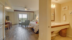 Une chambre d’hôtel comprenant un lit, un bureau avec une chaise, un balcon avec des rideaux, une salle de bain avec un lavabo et un miroir, et un ventilateur de plafond.