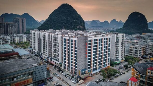 Un edificio residencial alto con varios pisos, rodeado de otros edificios y montañas al fondo.
