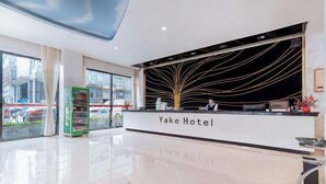 Zona de recepción de un hotel con diseño moderno, que incluye un mostrador de recepción, una máquina expendedora verde y grandes ventanales con vistas a la calle.
