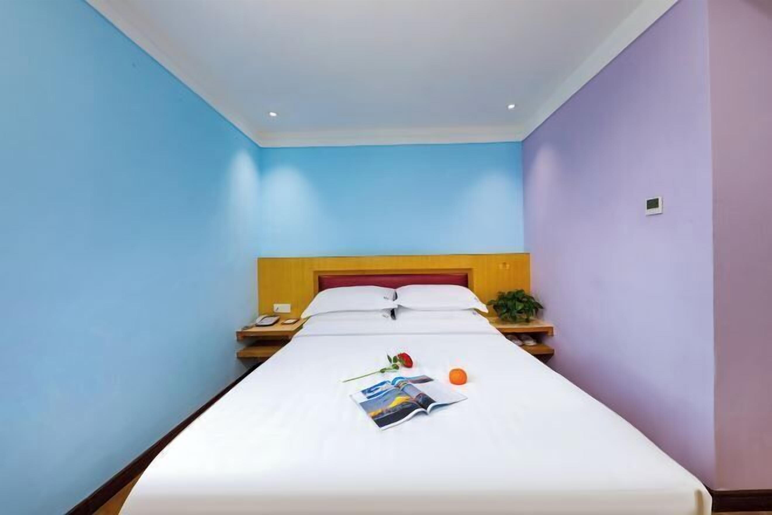 A hotel room with a bed, a nightstand, a phone, a magazine, a rose, and an orange.