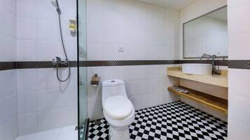 Casa de banho com cabine de chuveiro em vidro, sanita branca, lavatório com espelho e chão xadrez.