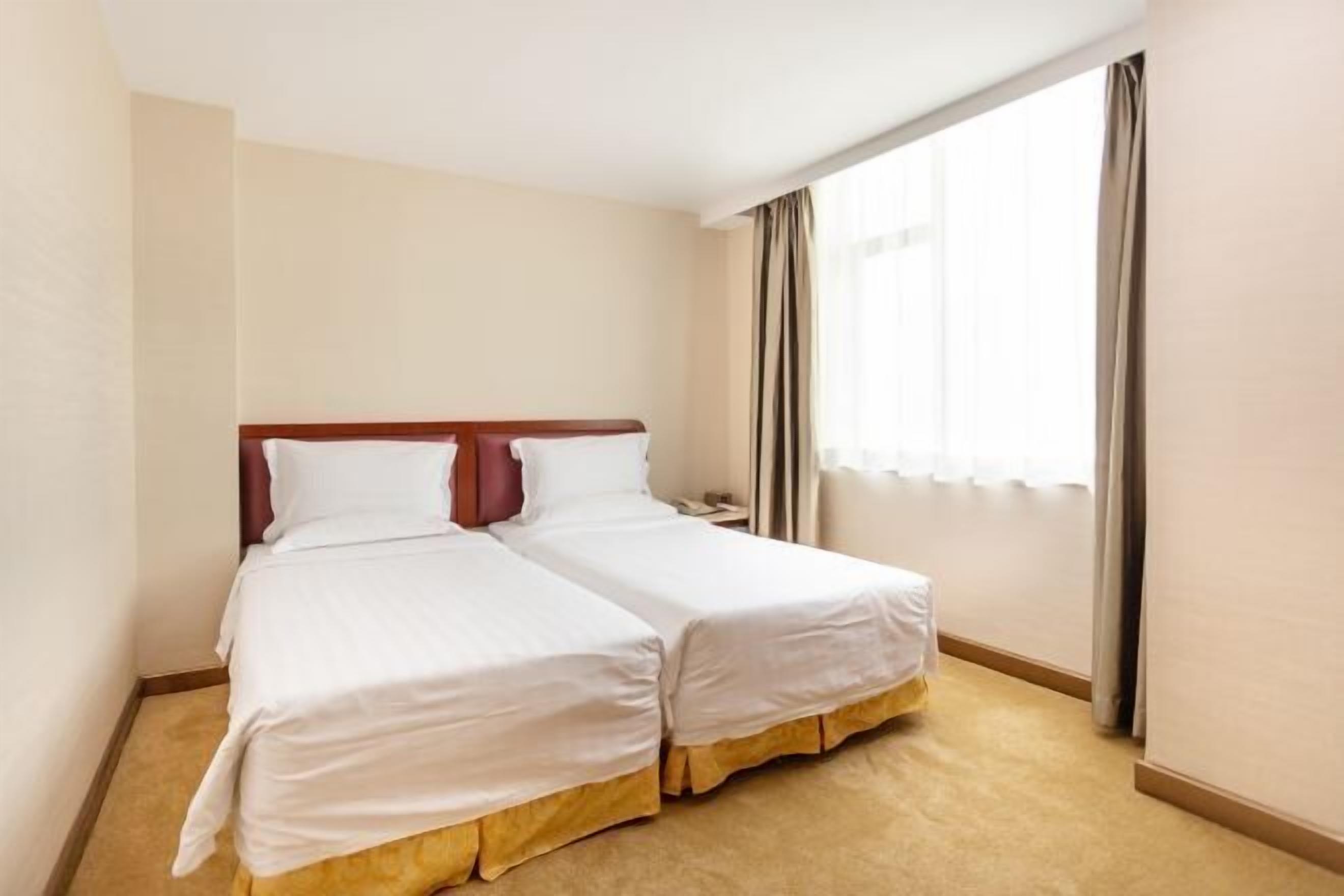 A hotel room with two single beds, a wooden headboard, beige walls, a large window with curtains, and a carpeted floor.