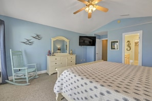 A bedroom with a bed, dresser, chair, ceiling fan, TV, and a door leading to another room.