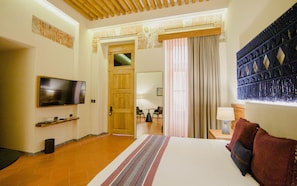 Quarto de hotel com uma cama grande, televisão, teto de madeira e vista para uma área de refeições.