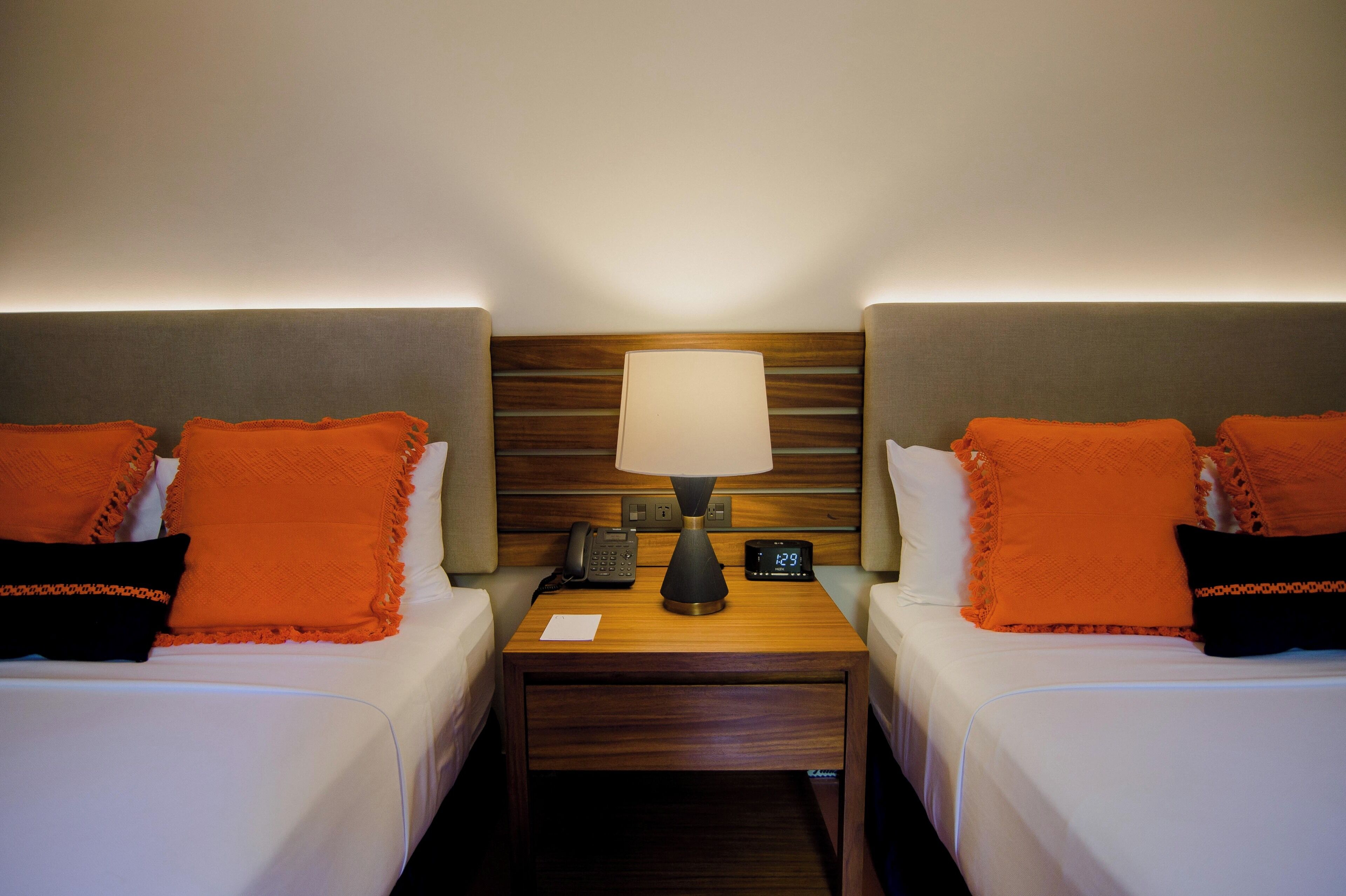 Habitación de hotel con dos camas, cada una con un cojín naranja y uno negro, una mesita de noche con una lámpara, un teléfono y un reloj digital.
