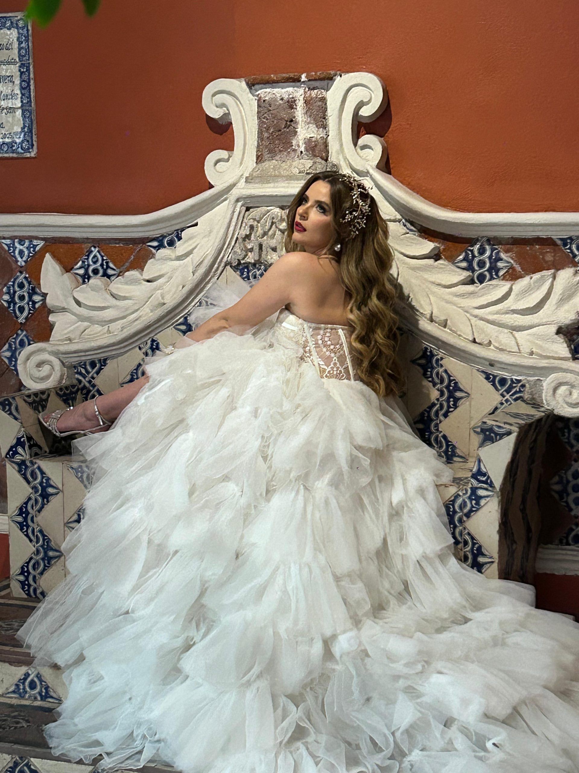 Une personne assise sur un banc doté d’un appui-tête décoratif, portant une robe de mariée blanche à la jupe volumineuse.