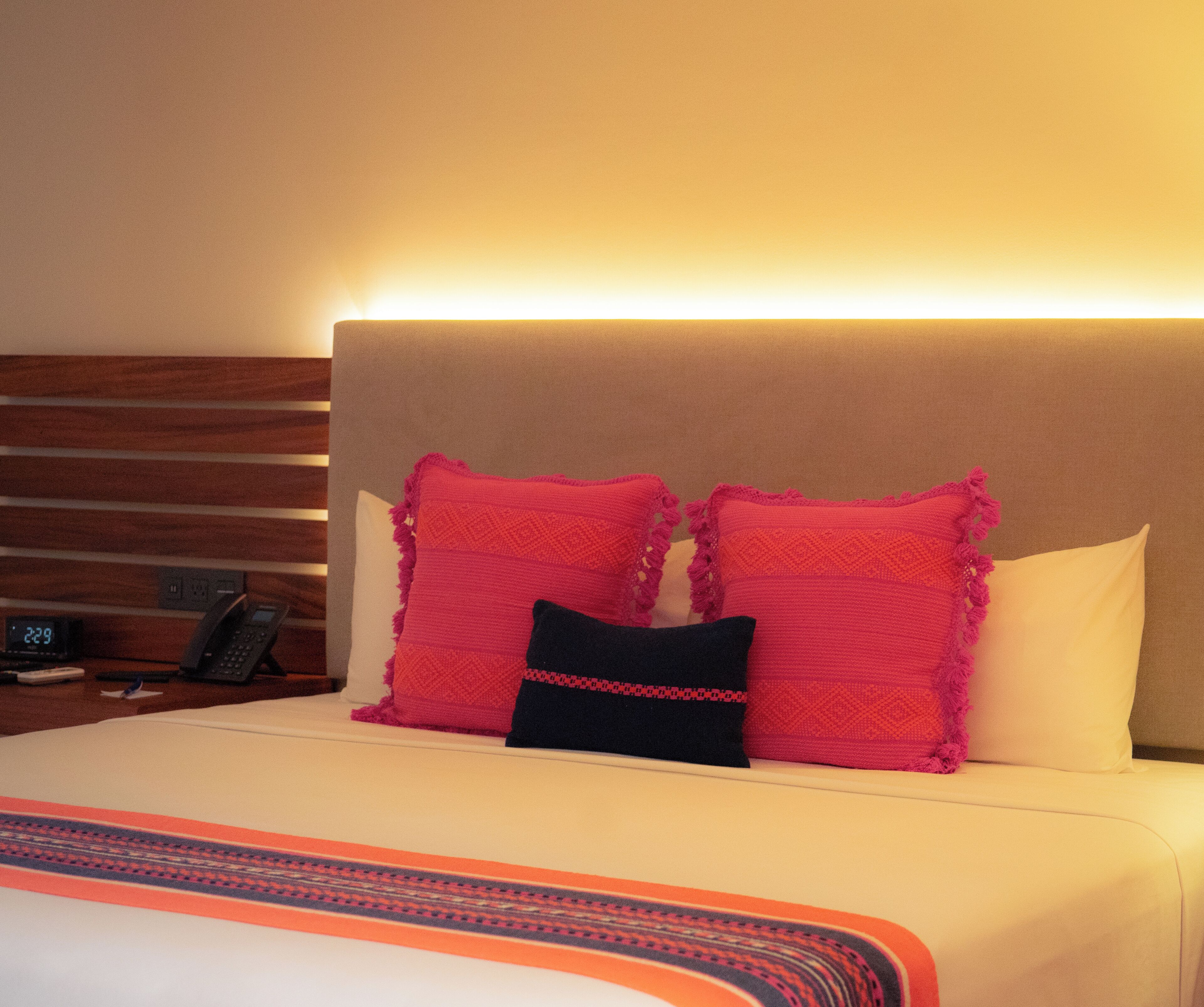 A hotel room with a bed, two pillows, a bedside table with a phone, and a wall clock.