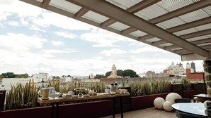 A rooftop terrace with a view of the city, featuring a table set with food and drinks, and a seating area with chairs and planters. - Casa Azulai Puebla Hotel Boutique (Puebla)
