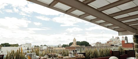 Una terraza en la azotea con vistas a la ciudad, que incluye una mesa con comida y bebidas, y un área de descanso con sillas y macetas.