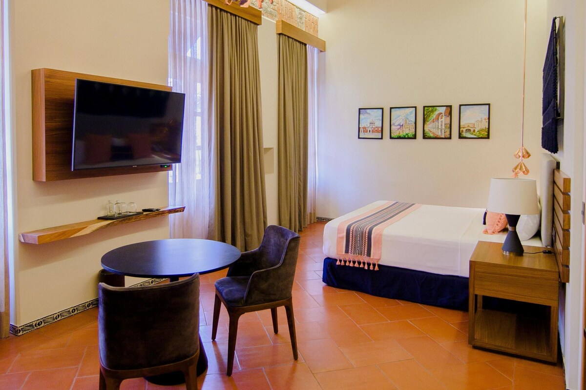 Habitación de hotel con cama, televisión montada en la pared, una mesita con sillas y una ventana con cortinas.