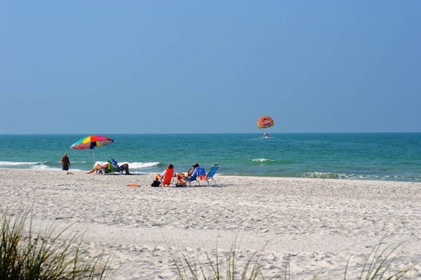 A beach with people relaxing under umbrellas, a parasail in the distance, and a clear blue sky.