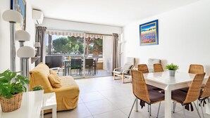 A bright, modern living room with a sofa, dining table, and chairs. There is a large window with curtains, a TV, and a painting on the wall. - Lovely apartment for 4 people with WIFI, A/C, TV and balcony (Le Lavandou)