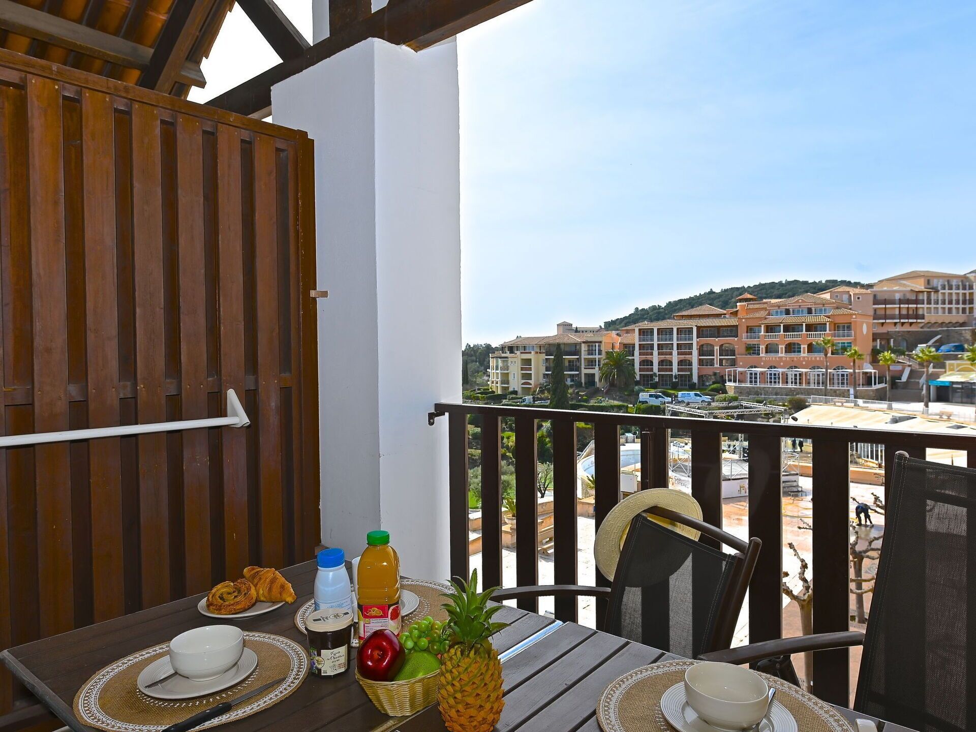 Een balkon met een ontbijttafel, compleet met croissants, fruit en drankjes, met uitzicht op een hotel en een glooiend landschap.