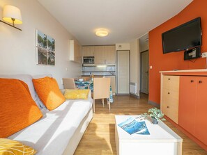 A compact living space with a sofa, a coffee table with magazines, a kitchenette, and a TV mounted on the wall.