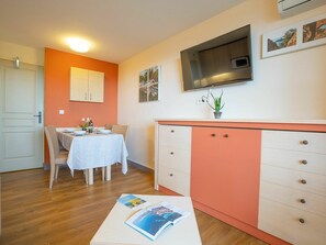A compact kitchen with a dining area, featuring a white table set for two, a cabinet with drawers, a microwave, and a flat-screen TV mounted on the wall.