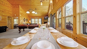 Interior de una cabaña de madera con mesa de billar, mesa de comedor para cuatro personas y amplias ventanas.