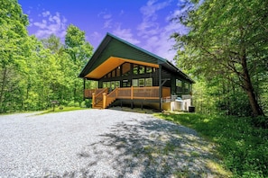 A lodge with a covered deck, a gravel driveway, and surrounded by dense forest.