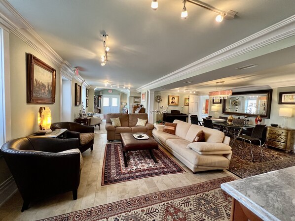 A spacious living room with a large sofa, armchairs, and a coffee table. There is a dining area with a table and chairs, and a kitchen in the background.