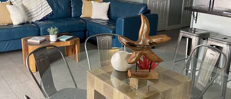 Ein Wohnzimmer mit einem Esstisch aus Glas, umgeben von Metallstühlen, einem blauen Sofa mit Kissen, einem Gemälde eines Piers und einem Deckenventilator.