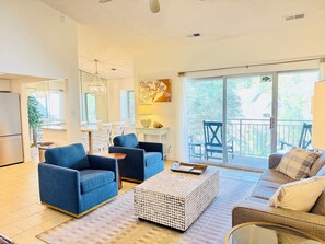 A spacious living area with a dining table, blue sofas, a coffee table, and a balcony with outdoor seating.