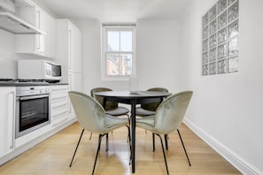 A modern kitchen with white cabinets, a stove, and a microwave. There is a dining table with four chairs and a window above it.