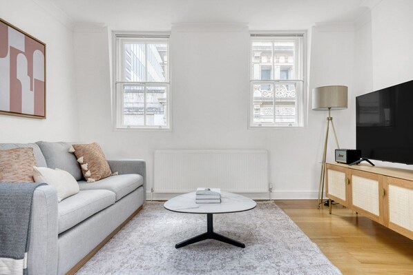A modern living room with a grey sofa, a round coffee table, a TV on a wooden cabinet, and large windows with a view of buildings. - Large Covent Garden 1BR, Charing Cross/Embankment station, by Blueground (London)
