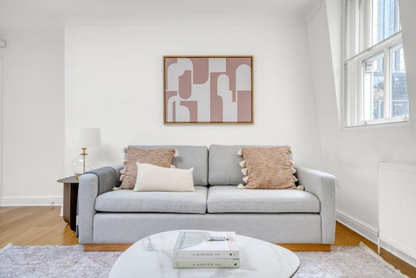 A living room with a grey sofa, two pillows, a round coffee table with books, a lamp, and a framed abstract artwork on the wall.