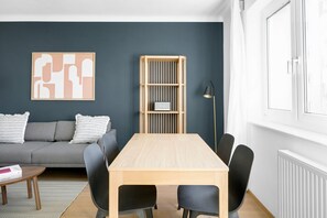 Una sala de estar moderna con un sofá gris, una mesa de centro de madera, una mesa de comedor con cuatro sillas, una estantería y una lámpara de pie.