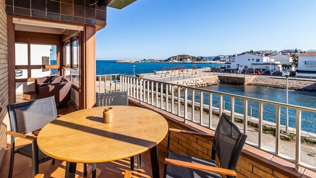 Un balcone con un tavolo e sedie di legno, affacciato su una zona costiera con un muro in pietra e edifici sullo sfondo.